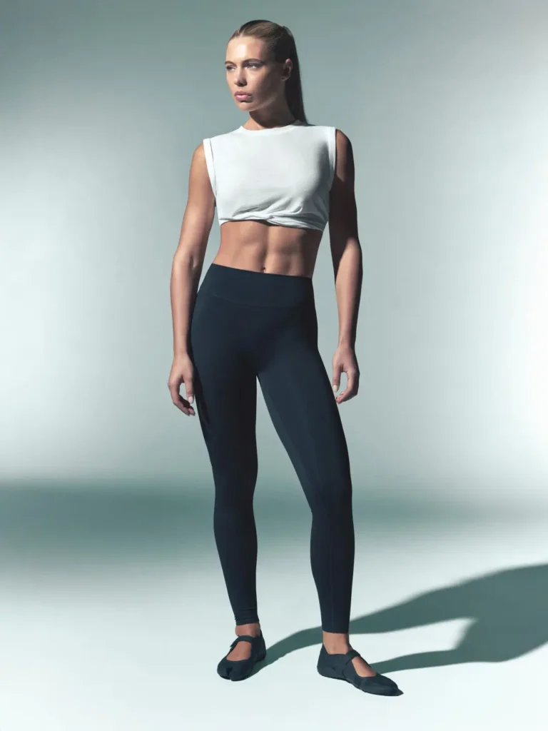 Woman standing in a white cropped sleeveless top, black leggings, and black split-toe flats against a pale studio backdrop