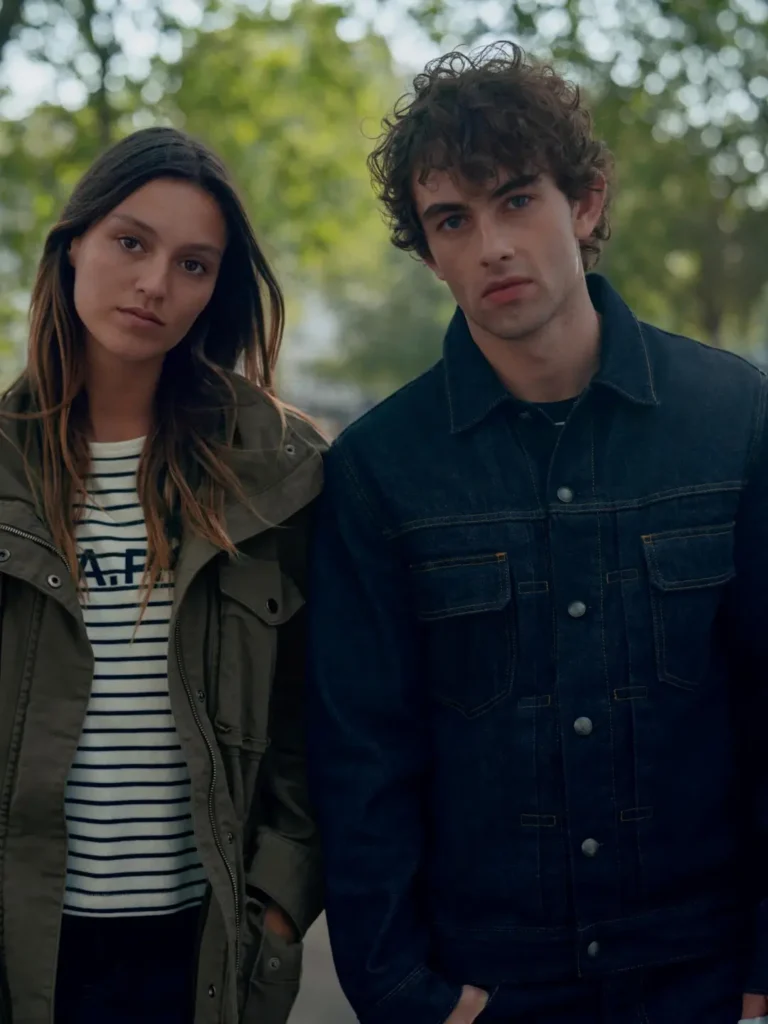 Two people stand outdoors; the woman wears an olive parka over a striped shirt, and the man wears a dark denim jacket