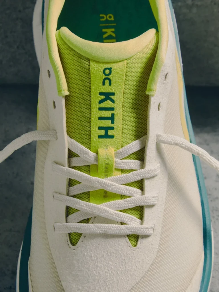 Close-up of a white sneaker with white laces, lime-green mesh tongue and teal trim on a dark floor