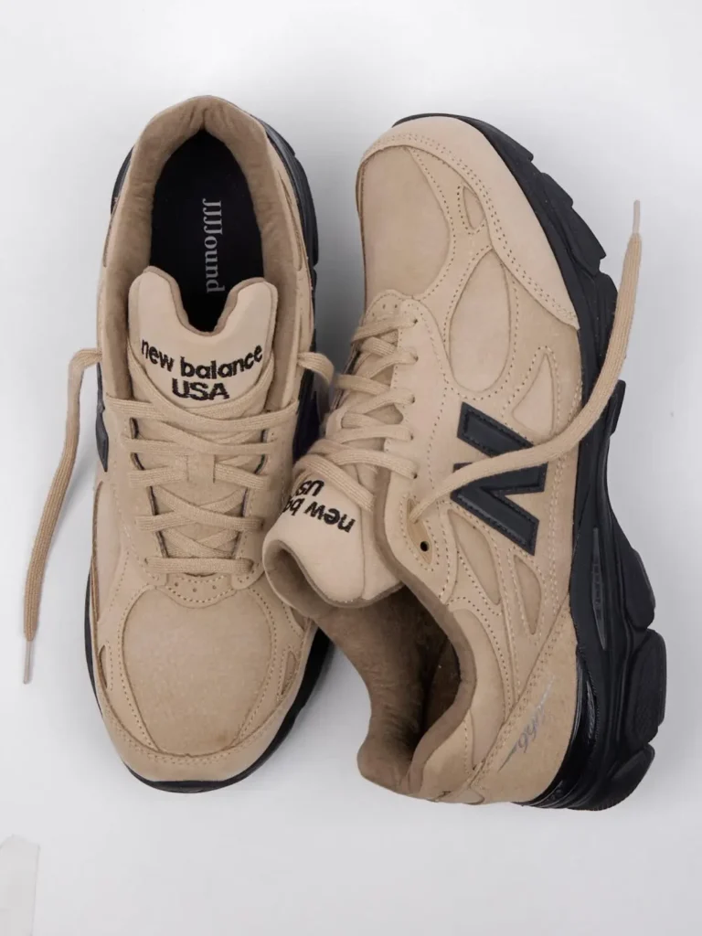 Pair of beige New Balance sneakers with black soles and oversized side logos, arranged on a white background