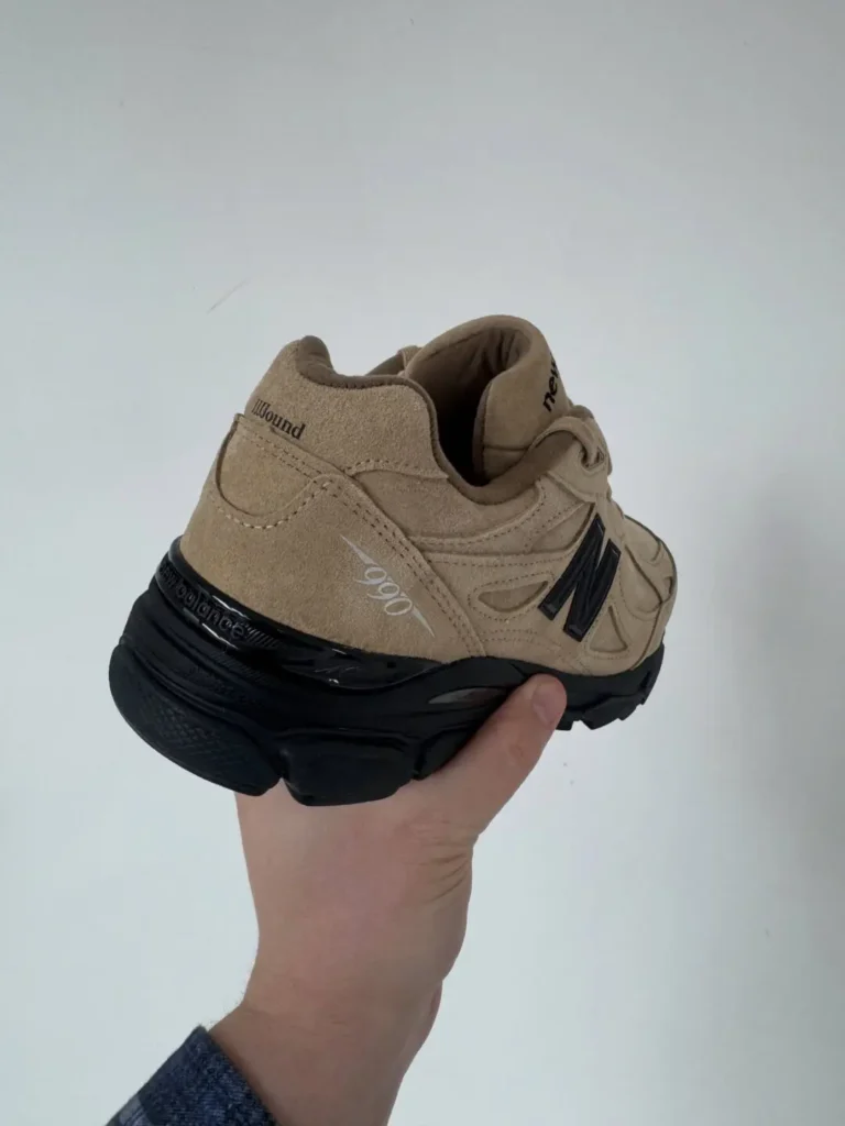 Hand holding a tan New Balance sneaker with a black sole against a light background