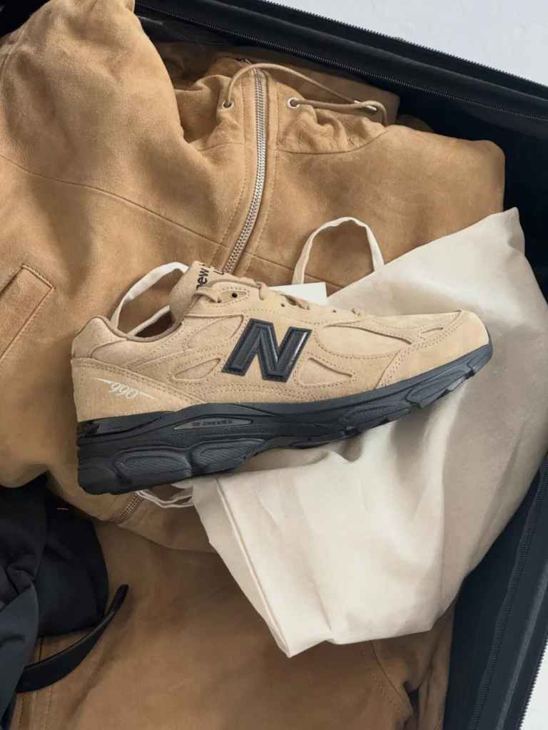 Beige New Balance sneaker resting on a tan jacket and white clothing inside an open suitcase