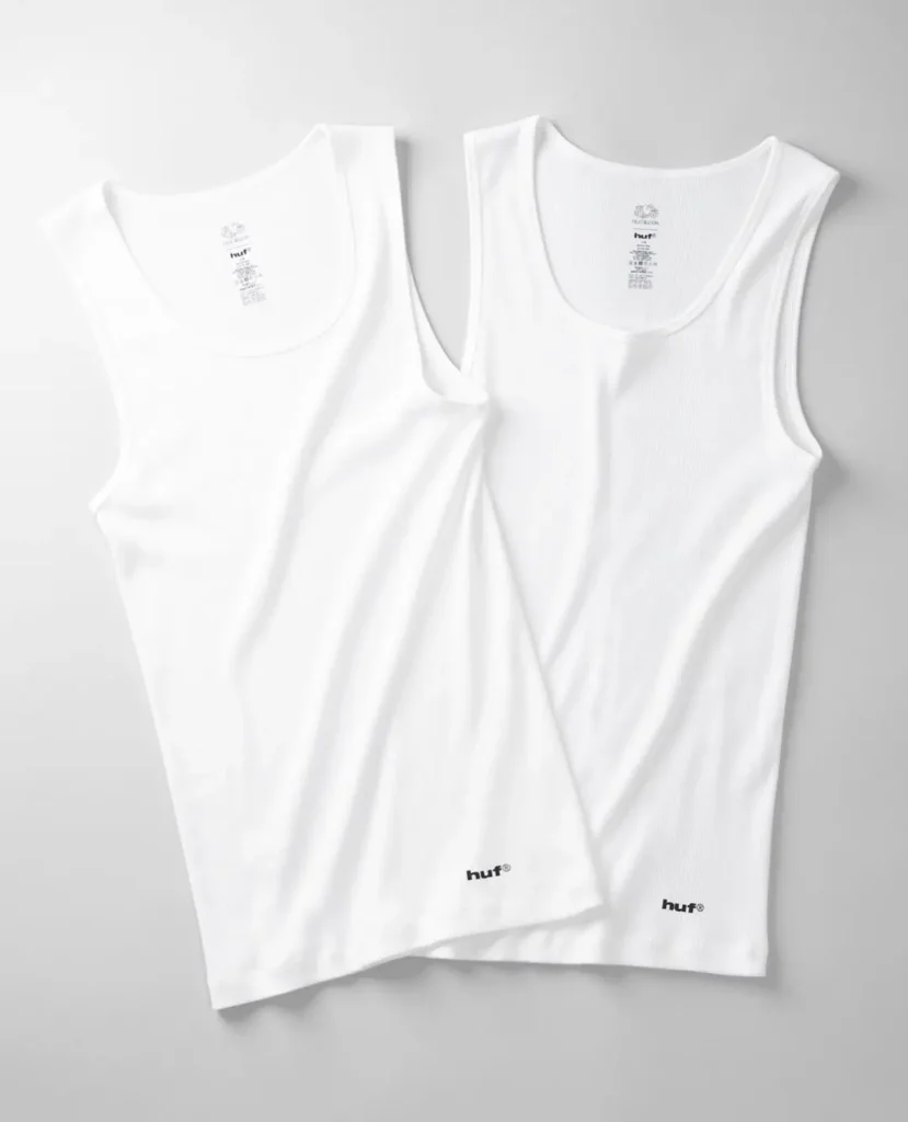 Two white HUF tank tops laid flat on a light gray surface, with small black logos near the hems