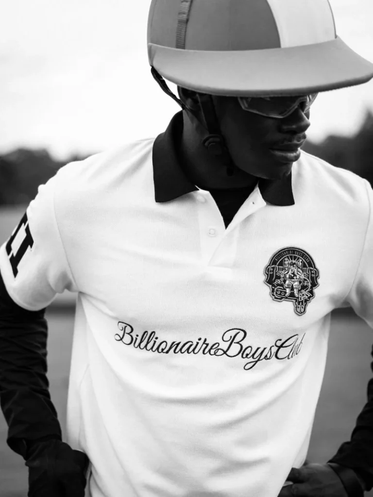 Black-and-white close-up of a person in a white cap and polo with a crest and “Billionaire Boys Club” script logo