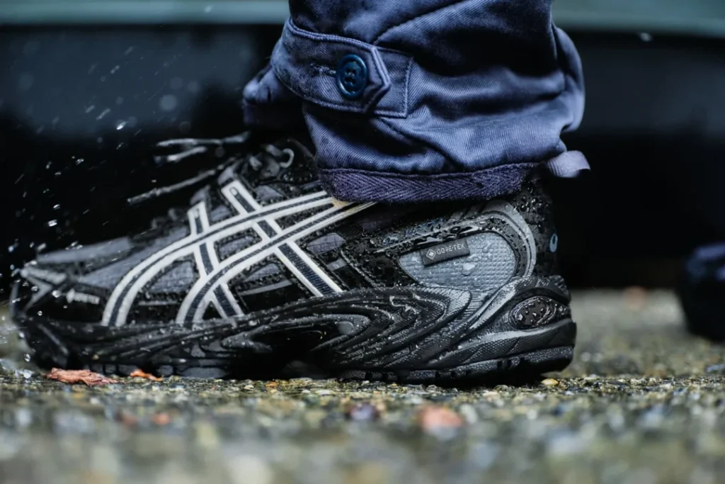 Close-up of a wet black ASICS sneaker with white stripes, worn with navy pants, standing on wet pavement
