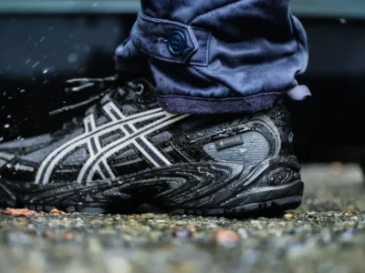 Close-up of a wet black ASICS sneaker with white stripes, worn with navy pants, standing on wet pavement