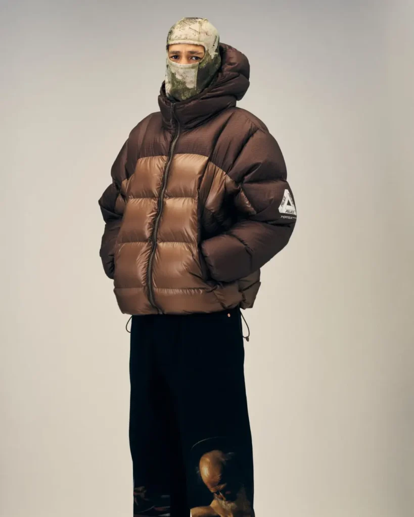 Person in a camouflage face covering and brown-and-black puffer jacket, hands in pockets, against a plain beige background