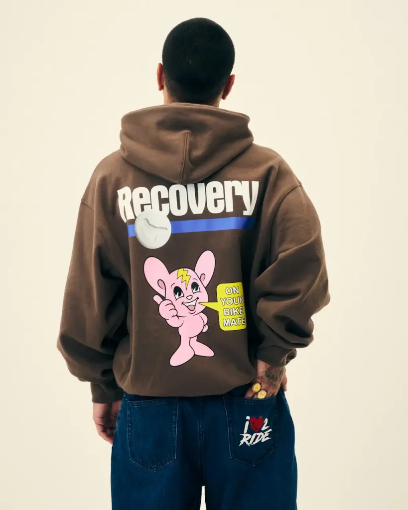 Rear view of a person in a brown hoodie reading “RECOVERY,” with a cartoon graphic, blue jeans, and a hand in the pocket