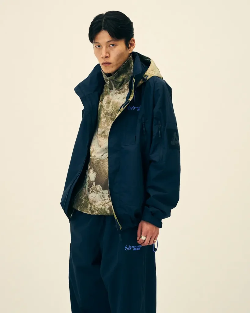 Model wearing a navy hooded jacket and pants over a mottled beige fleece, standing against a plain cream background