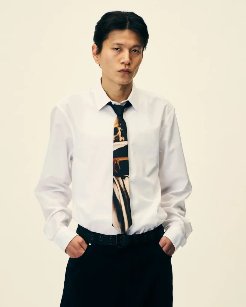 Model stands against a beige background wearing a white shirt, printed black tie, black trousers, and wide grommet belt