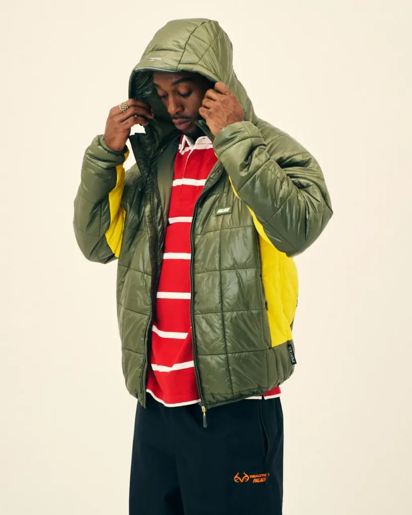 Person adjusts the hood of a quilted olive-green jacket with yellow lining over a red-and-white striped shirt against a