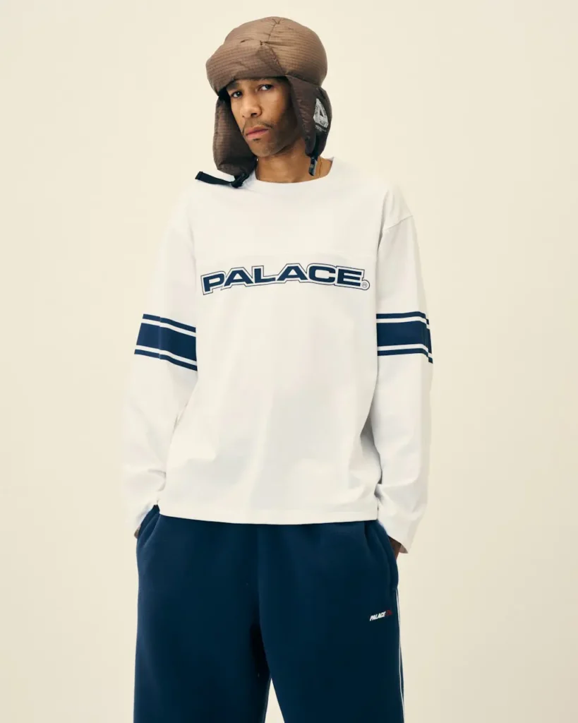 Model wearing a brown padded trapper hat, white Palace long-sleeve shirt, and navy track pants against a cream backdrop
