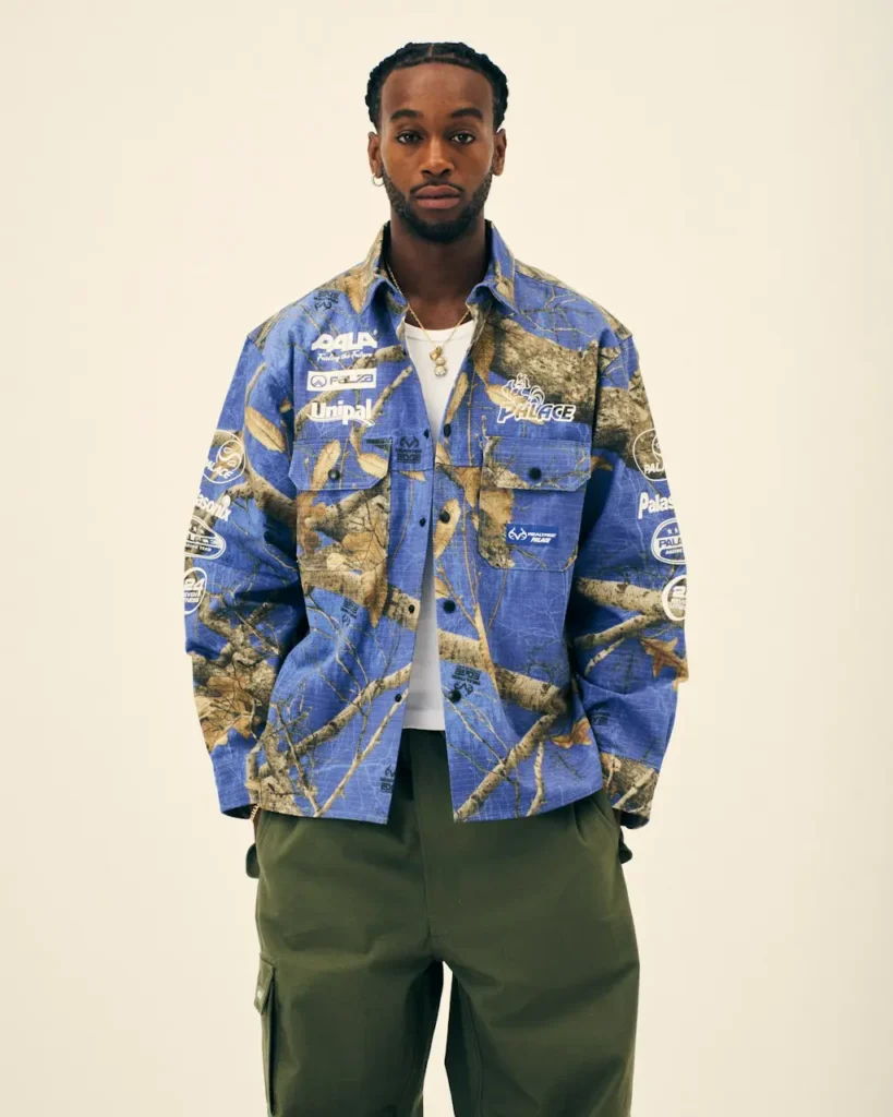 Man wears a blue and tan graphic Palace overshirt over a white shirt and olive cargo pants against a plain beige backdrop
