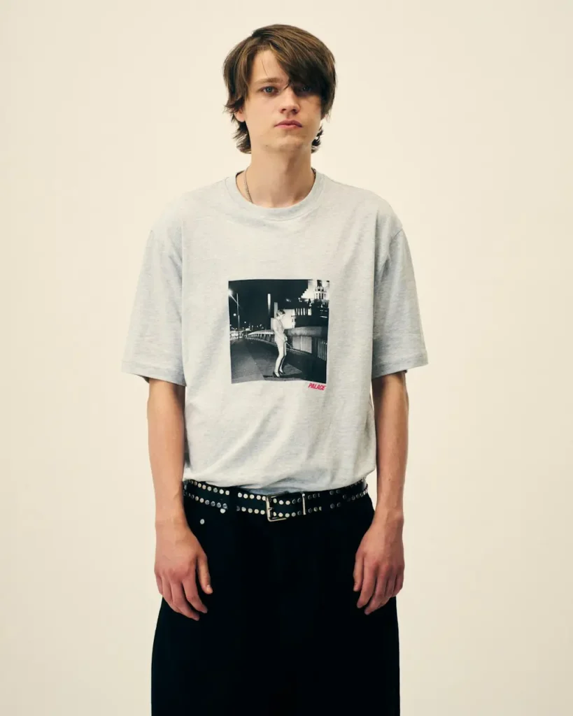 Model stands against a beige backdrop wearing a gray graphic T-shirt, black studded belt, and black trousers