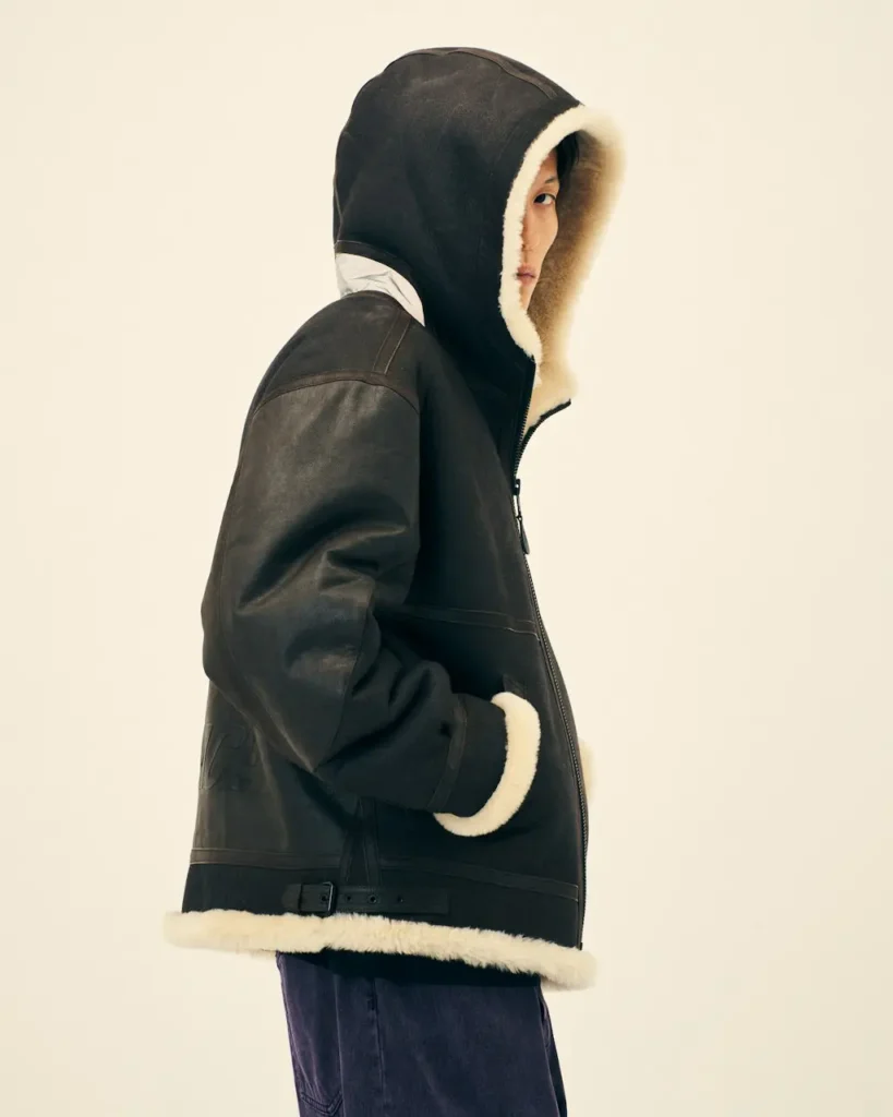 Person in profile wearing a black leather shearling-lined hooded jacket, hands in pockets, against a cream background