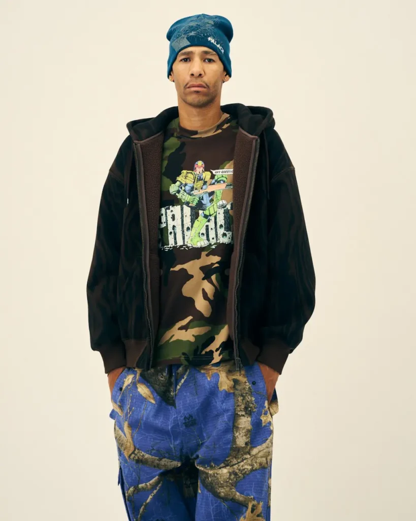 Model stands against a plain cream background wearing a blue Palace beanie, black jacket, camouflage Judge Dredd shirt and