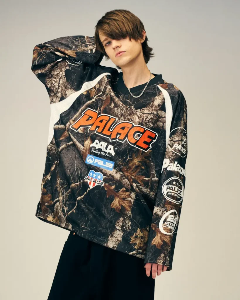 Model posing in a dark woodland-print Palace jersey with orange logo and sleeve patches against a plain beige background