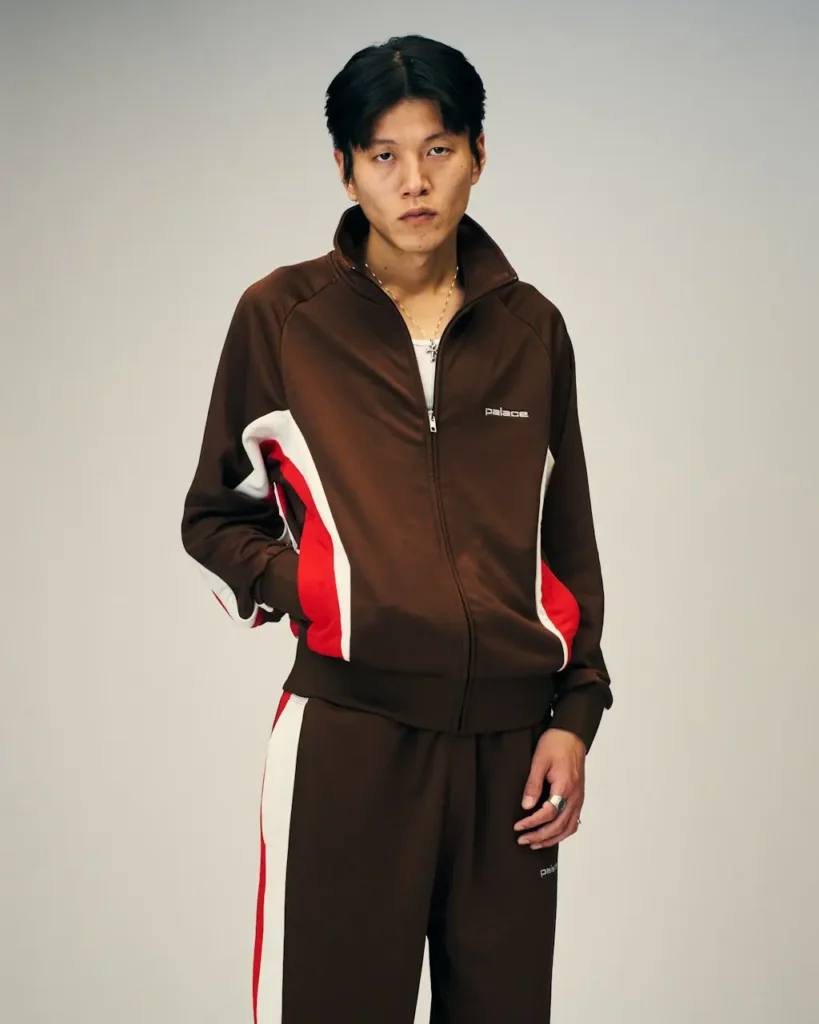 Model stands against a plain background in a brown Palace tracksuit with red and white side panels, one hand in a pocket