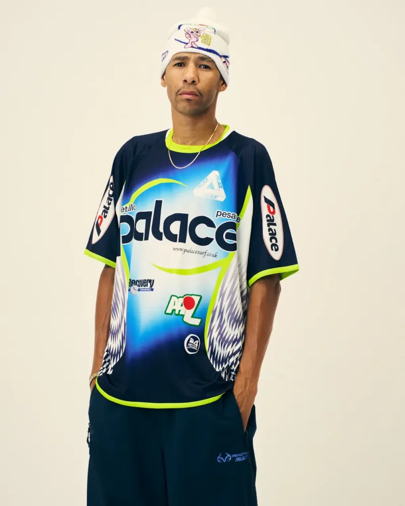 Man stands against a cream backdrop wearing a navy Palace racing jersey with neon trim, logos and matching pants, plus a