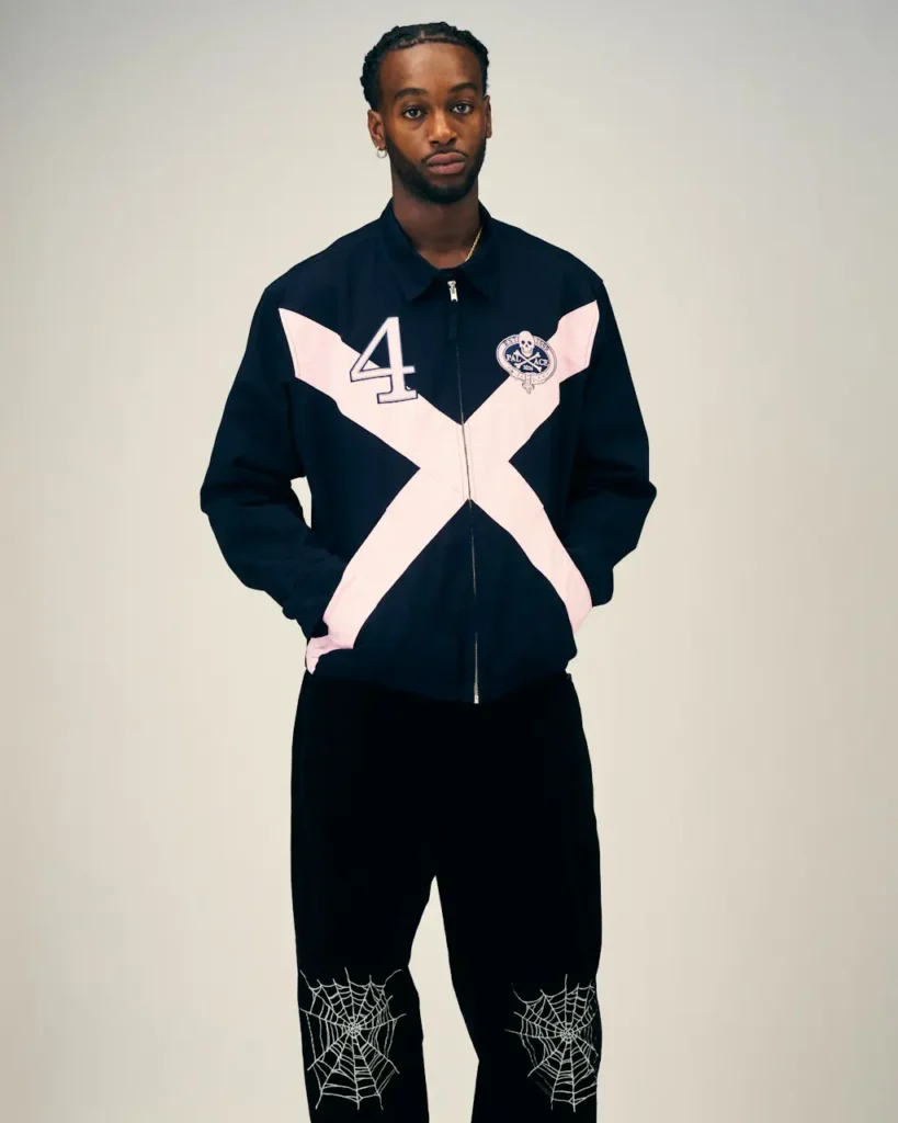 Man stands against a plain beige backdrop wearing a navy jacket with a pale pink X, number 4 and crest, plus black pants