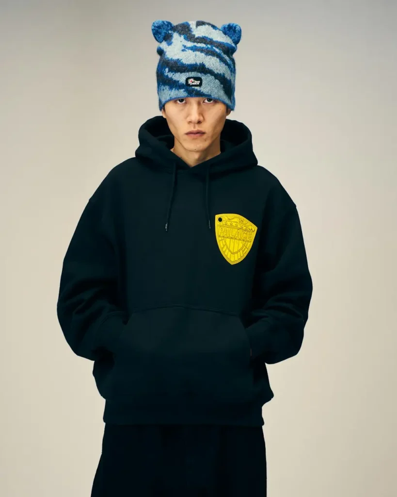 Model stands against a beige backdrop wearing a black hoodie with a yellow shield patch and blue patterned bear-ear beanie