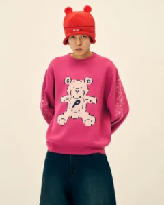 Model wearing a red pom-pom hat and pink sweater with a large bear graphic, posed against a beige background