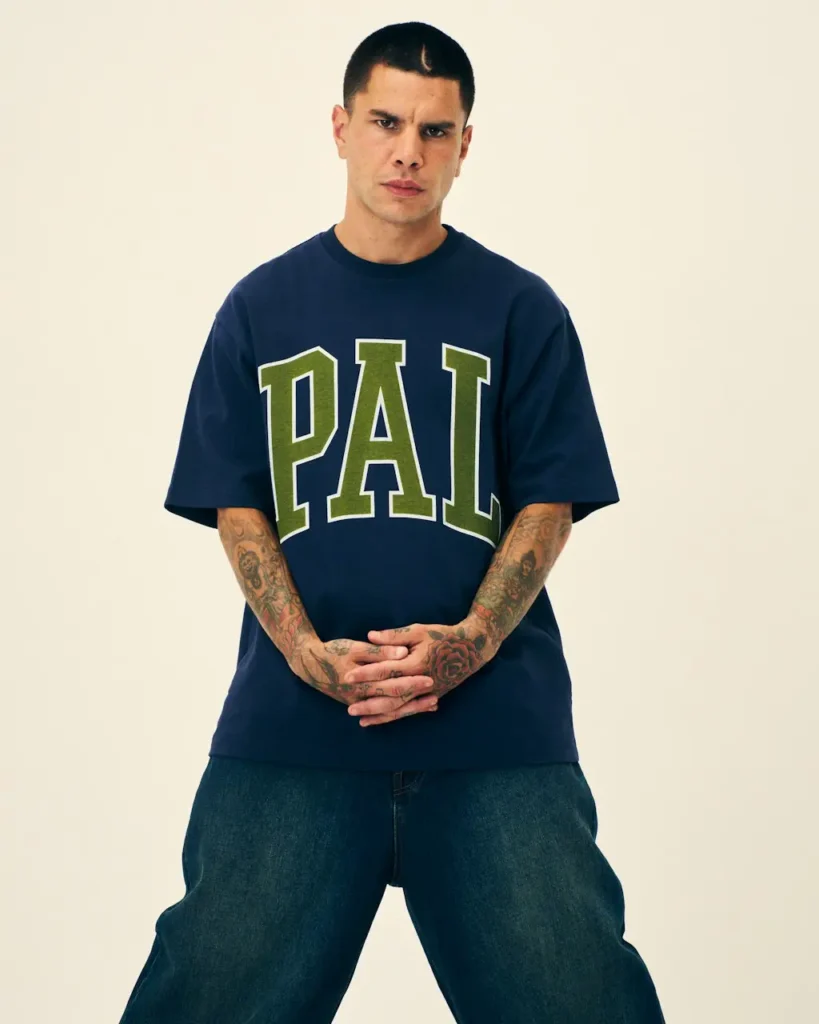 Tattooed man stands with hands clasped, wearing a navy T-shirt with large olive PAL lettering and baggy jeans against a