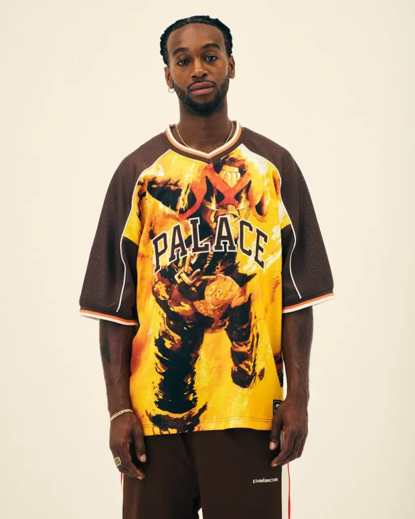 Man standing against a plain cream background wearing a yellow-and-brown Palace graphic jersey and black track pants