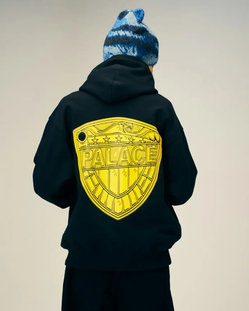 Person seen from behind wearing a black hoodie with a large yellow Palace shield graphic and a blue patterned knit beanie