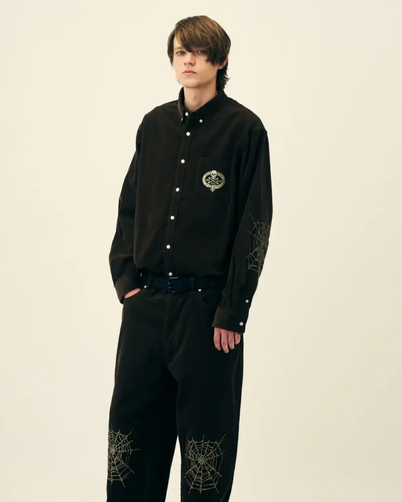 Model wearing a black Palace shirt and trousers with web graphics on the sleeve and knees, posed against a cream backdrop