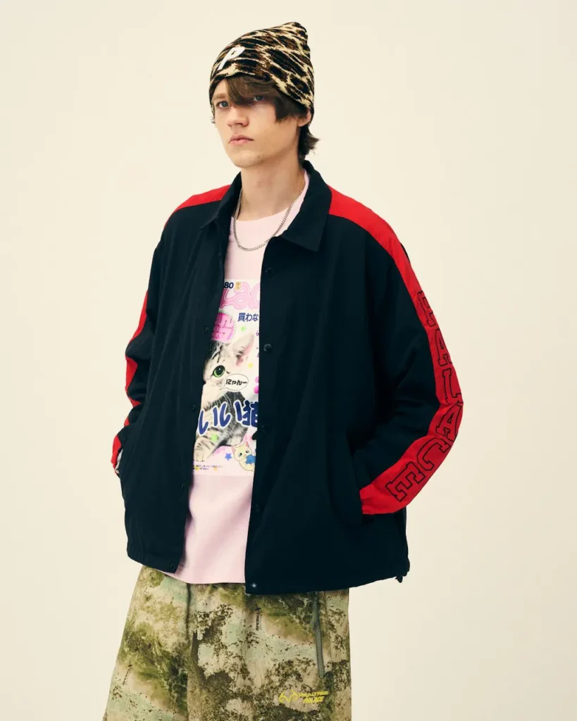 Model stands with hands in pockets wearing a black jacket with red sleeves, pink graphic shirt, camo pants and animal-print