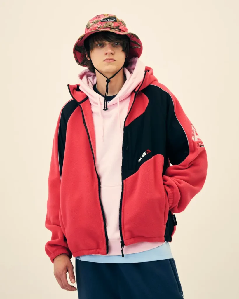 Model wearing a red and black Palace jacket, pink hoodie, and patterned bucket hat against a beige background