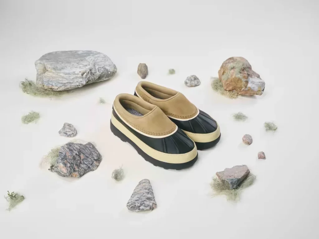 Pair of tan-and-black waterproof mule shoes arranged among rocks and tufts of moss on a white background