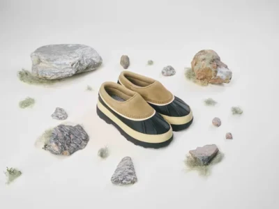 Pair of tan-and-black waterproof mule shoes arranged among rocks and tufts of moss on a white background
