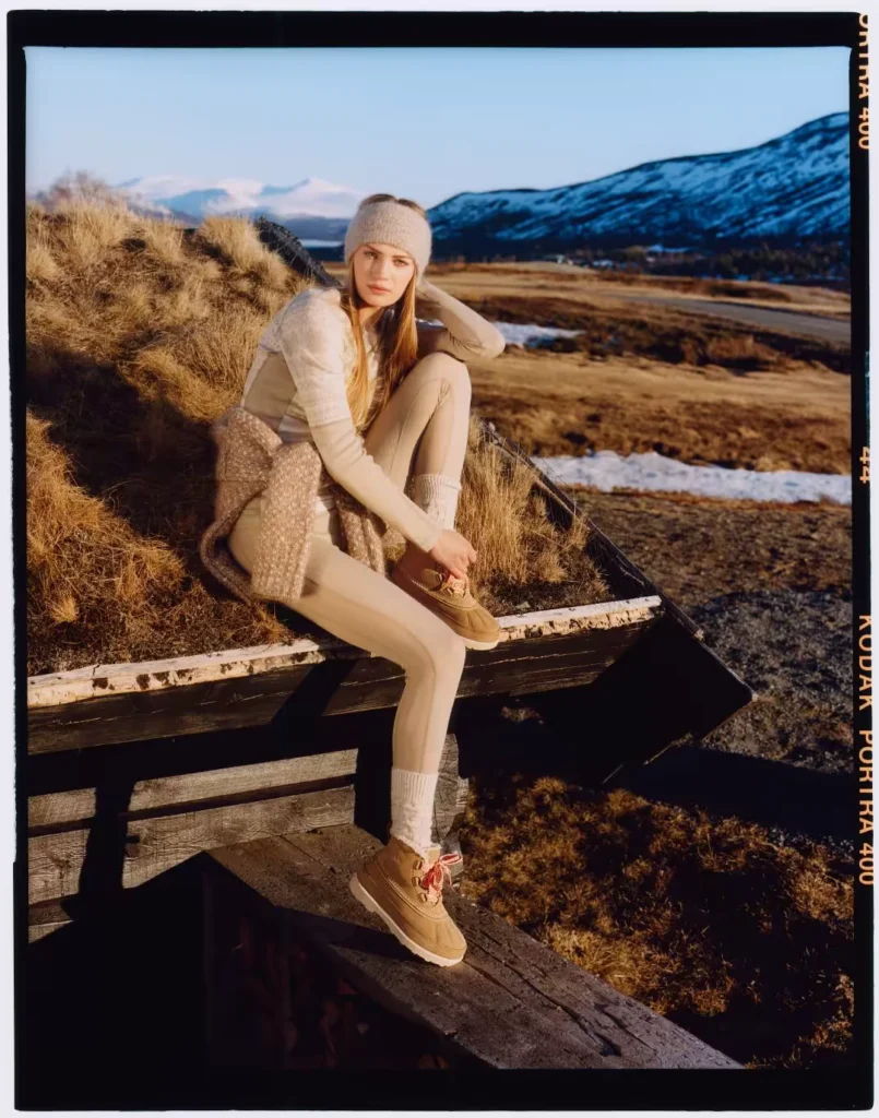 Model in beige knitwear and leggings sits on a wooden fence, wearing tan lace-up mules, with snowy mountains behind her