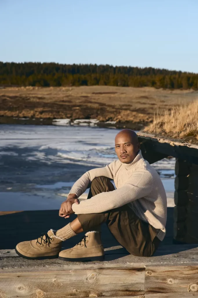 Bald man in a cream sweater sits by a frozen lake, wearing tan lace-up boots and brown pants