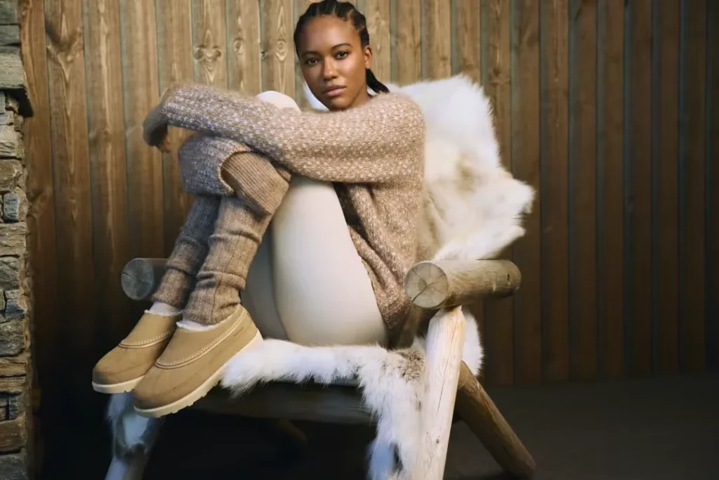 Woman seated on a fur-covered wooden chair, wearing a beige knit sweater, leg warmers, and tan waterproof mule boots
