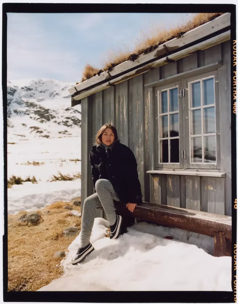 Person in a black jacket and gray leggings sits outside a weathered cabin in snow, wearing black-and-white mules