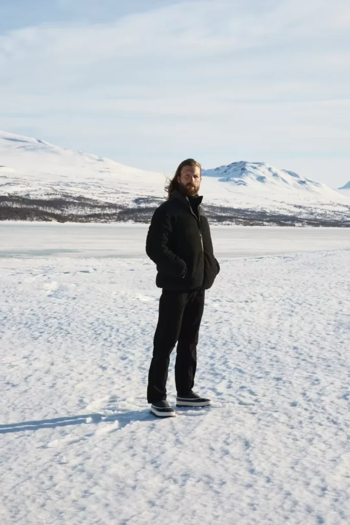 Bearded man in black clothing and mules stands on a snowy frozen lake with mountains behind him