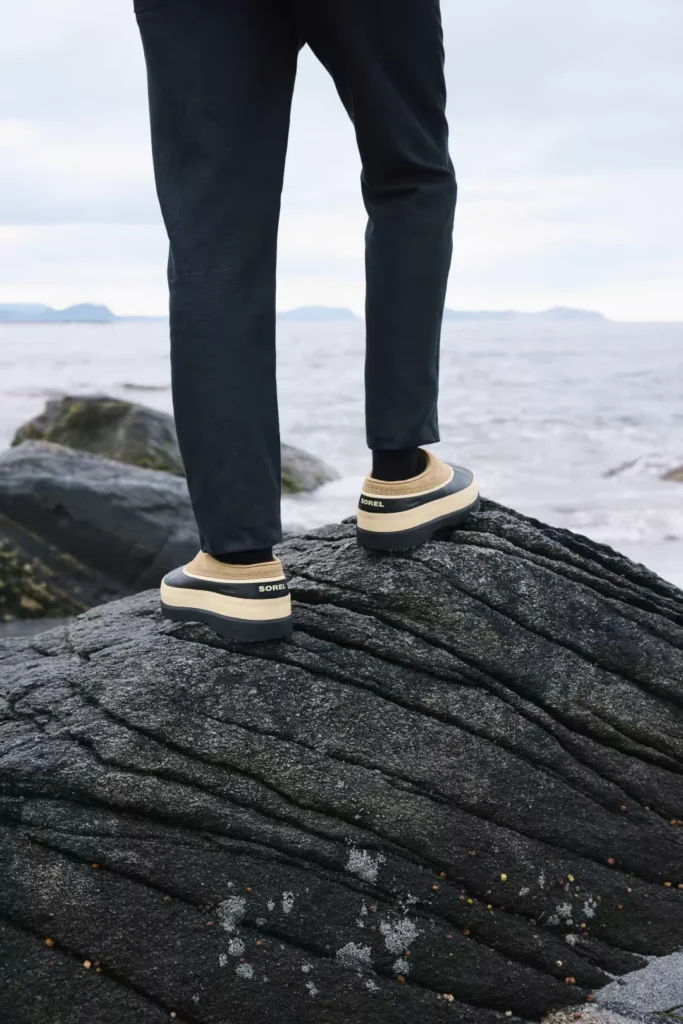 Person in black pants wearing cream-and-black SOREL mules, standing on dark coastal rocks beside the sea