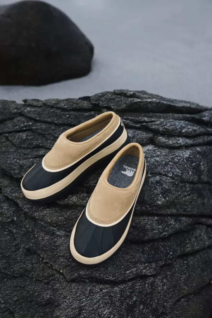 Two tan and black Sorel mules rest on dark textured fabric, with a black rock in the background