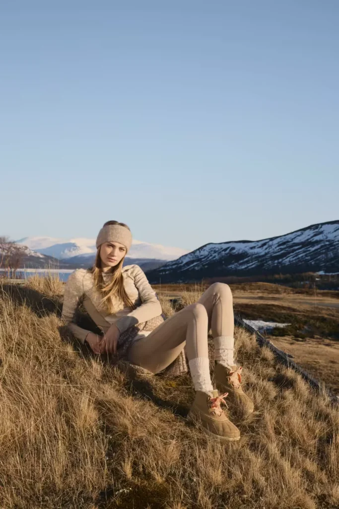 Woman seated in dry grass wearing beige clothing and lace-up boots, with snowy mountains and a lake behind her
