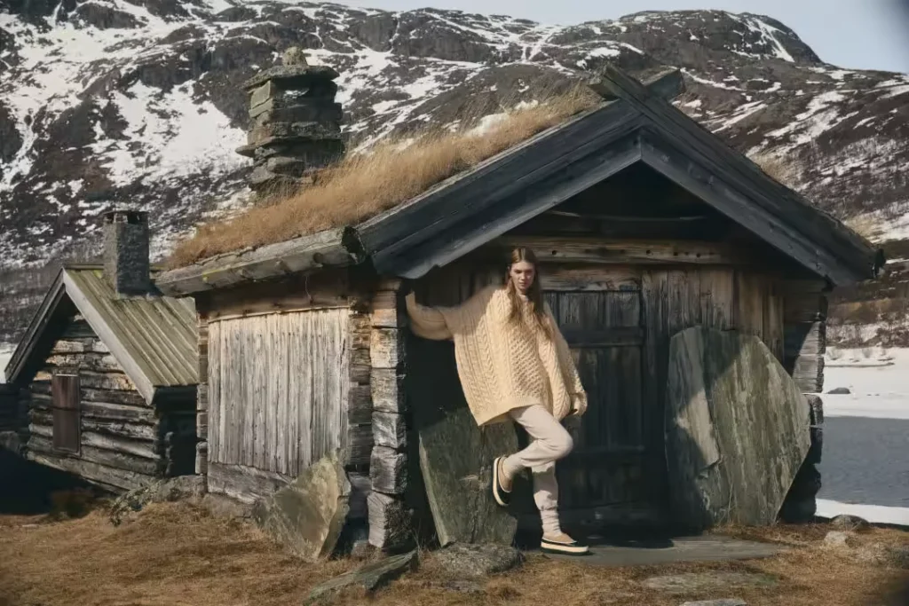 Model in a cream cable-knit sweater poses in beige waterproof mules outside a rustic cabin with snowy mountains beyond