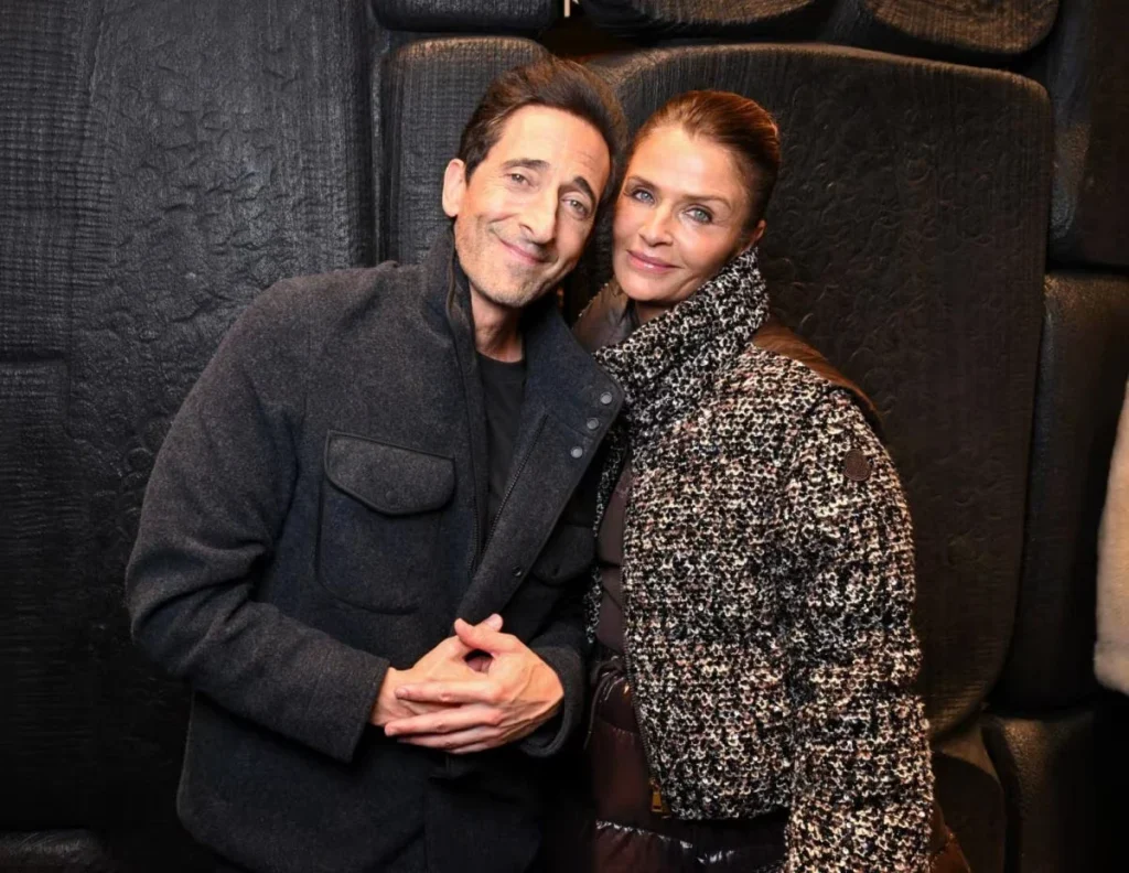 Adrien Brody and Georgina Chapman pose together against a dark textured backdrop; she wears a speckled tweed jacket and scarf