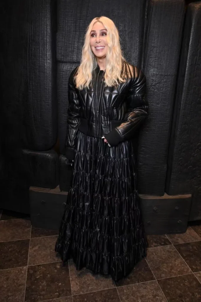 Cher poses in a black quilt leather jacket and glossy tiered floor-length skirt against dark curtains