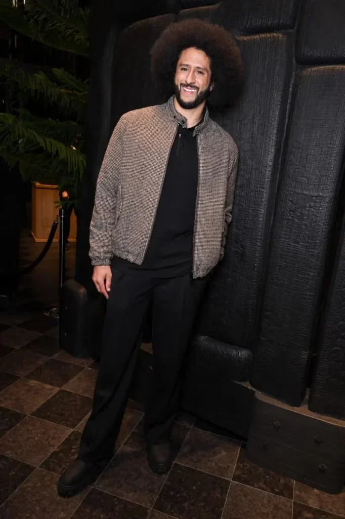 A smiling man with an afro wears a brown checked jacket over black clothing, posing before a dark textured backdrop