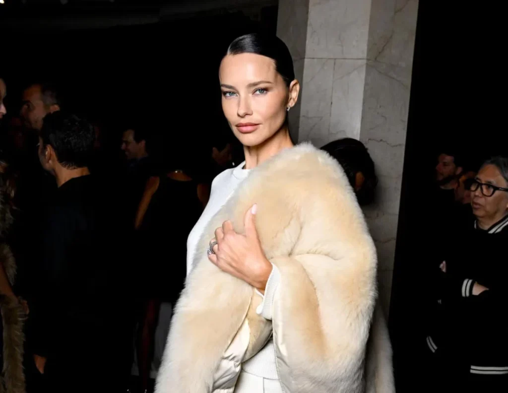 A woman poses indoors, holding a cream-colored fur coat over a white outfit as guests gather behind her