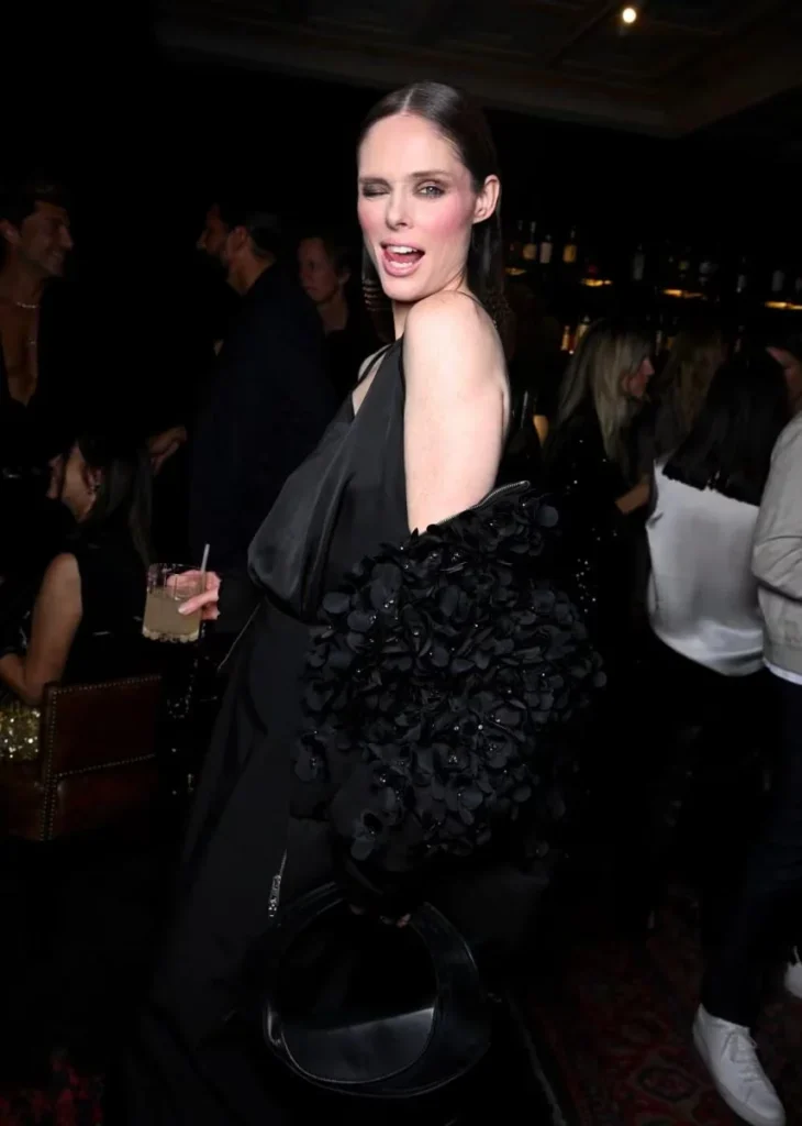 A woman wink while holding a drink in a black gown with a floral-textured jacket at a crowded event