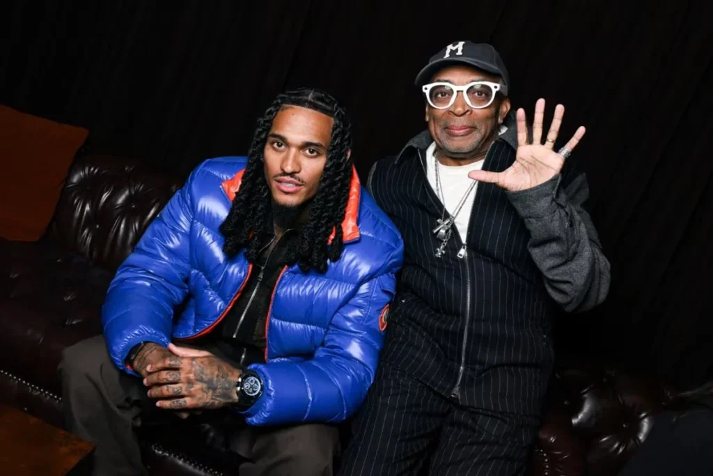 Left one man as right Spike Lee sit on a dark leather sofa; one wears a bright blue puffer jacket, while the other waves in white glasses and a cap