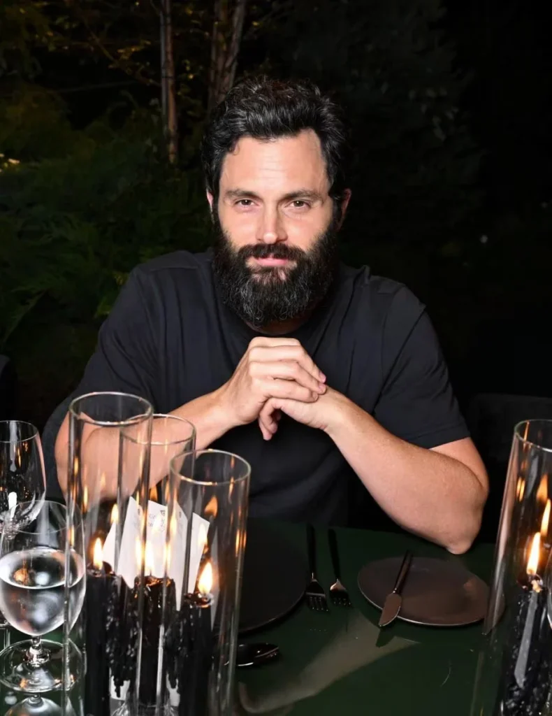 Bearded man in a black shirt sits with clasped hands at a candlelit dinner table surrounded by greenery
